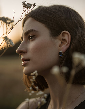 Woman with dark hair wearing sapphire gemstone earring, standing among wildflowers at sunset, soft natural light and warm tones